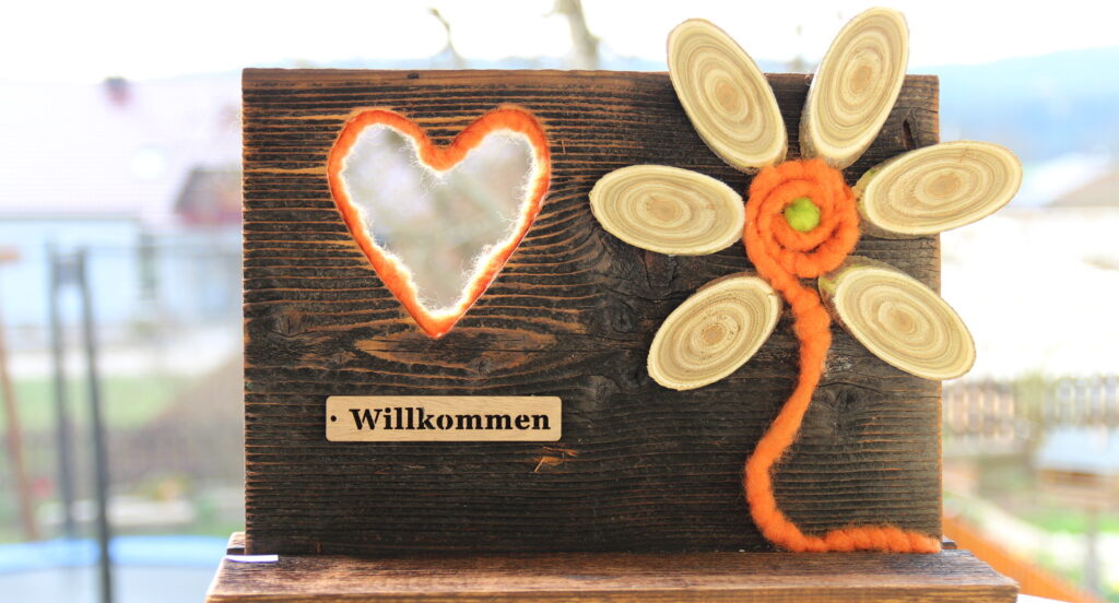 Holzschild mit Herzausschnitt und Aufschrift "Willkommen" und eine Blume aus Filz und Holzscheiben als Blütenblätter