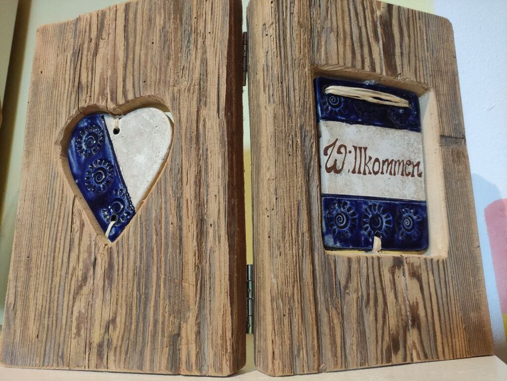 klappbares Holzbuch mit blau-weißem Tonschild 'Willkommen' auf Vorderseite und blau-weißem Tonherz auf Rückseite