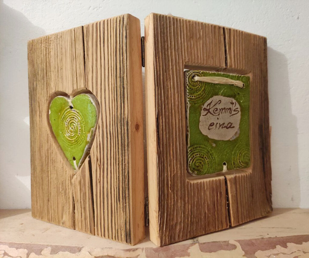 klappbares Holzbuch mit grünem Tonschild 'Kemmt's eina' auf Vorderseite und grünem Tonherz auf Rückseite